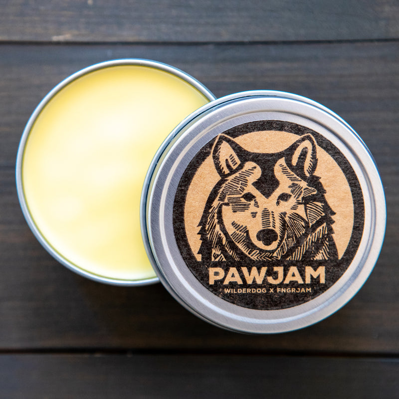 Open tin of PAWJAM all-natural salve, creamy yellow, wolf graphic on lid, on a dark wooden surface.